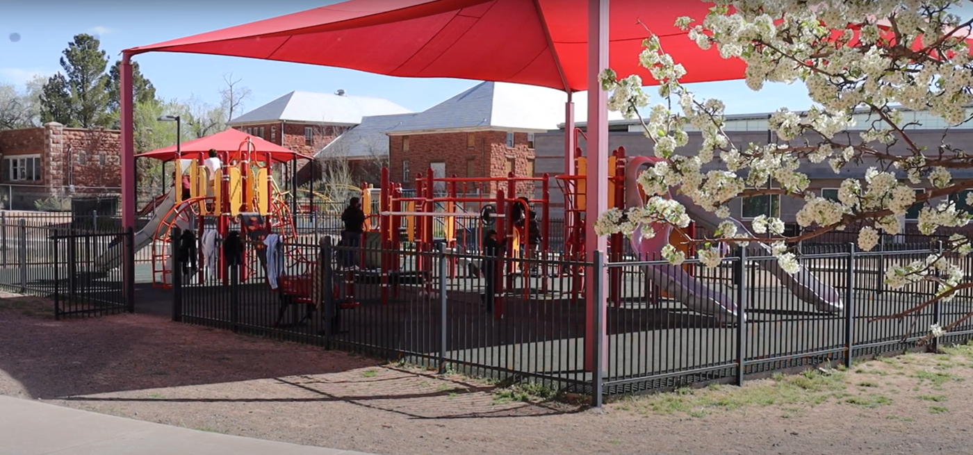 A bright red umbrella stands in front of a colorful playground filled with swings and slides.