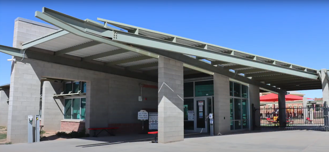 Front entrance of Tuba City Boarding school featuring a sloped roof and double doors, surrounded by a small pathway and greenery.